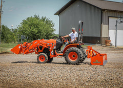 A man driving a Kubota compact tractor with a front loader and a Land Pride BB12 series box scraper across a gravel lot by a steel building